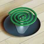 A green mosquito repellent coil in a spiral shape sits on a metal stand placed on a dark round plate, which rests on a light wooden floor—ready to ward off pests from your yard.