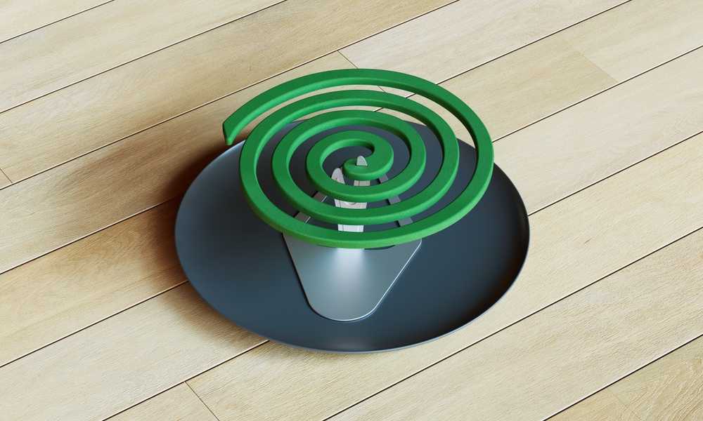 A green mosquito repellent coil in a spiral shape sits on a metal stand placed on a dark round plate, which rests on a light wooden floor—ready to ward off pests from your yard.