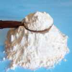 A pile of white powder, possibly boric acid used to get rid of roaches, sits on a blue surface with a wooden spoon decorated with blue and red beads resting on top.