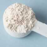 A white plastic scoop filled with fine, off-white diatomaceous earth sits on a light blue surface, ready to be used for natural flea control.