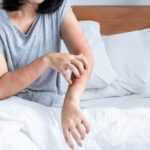 A person sitting on a bed scratches a red, irritated rash on their left forearm—possible symptoms of dust mites. They wear a gray T-shirt and white bedding is visible as they try to protect themselves. Their face is partially out of frame.