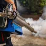 A person operates a fogging machine emitting white smoke onto a concrete surface for pest control, helping to maintain a pest-free home, with greenery visible in the background.