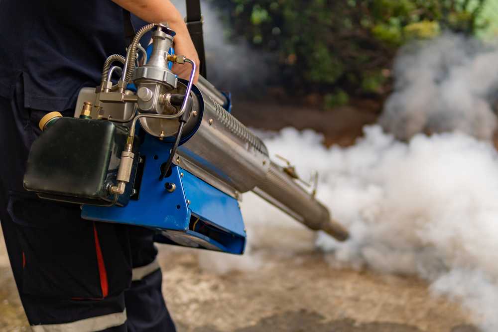 A person operates a fogging machine emitting white smoke onto a concrete surface for pest control, helping to maintain a pest-free home, with greenery visible in the background.