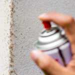 A hand sprays insecticide onto a corner of a wall covered with many small ants. The focus is on the spray can and the ants crawling on the textured surface.