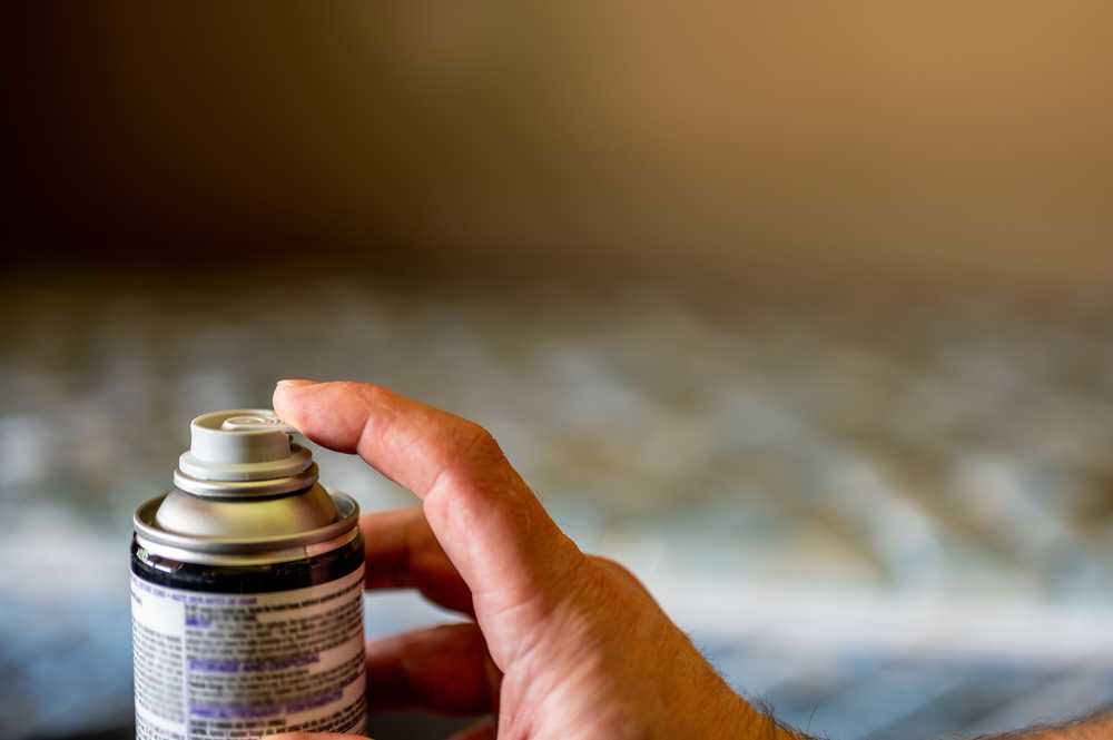 A close-up of a hand pressing the nozzle of an aerosol spray can, with a blurred background. The can has a label with text, but the contents are not identifiable.
