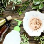 A bowl filled with liquid and many small flies sits beside a wine bottle, a pump bottle, and green leafy branches on a wooden surface. The scene shows a homemade fruit fly trap, much like a safe ant killer for natural pest control at home.