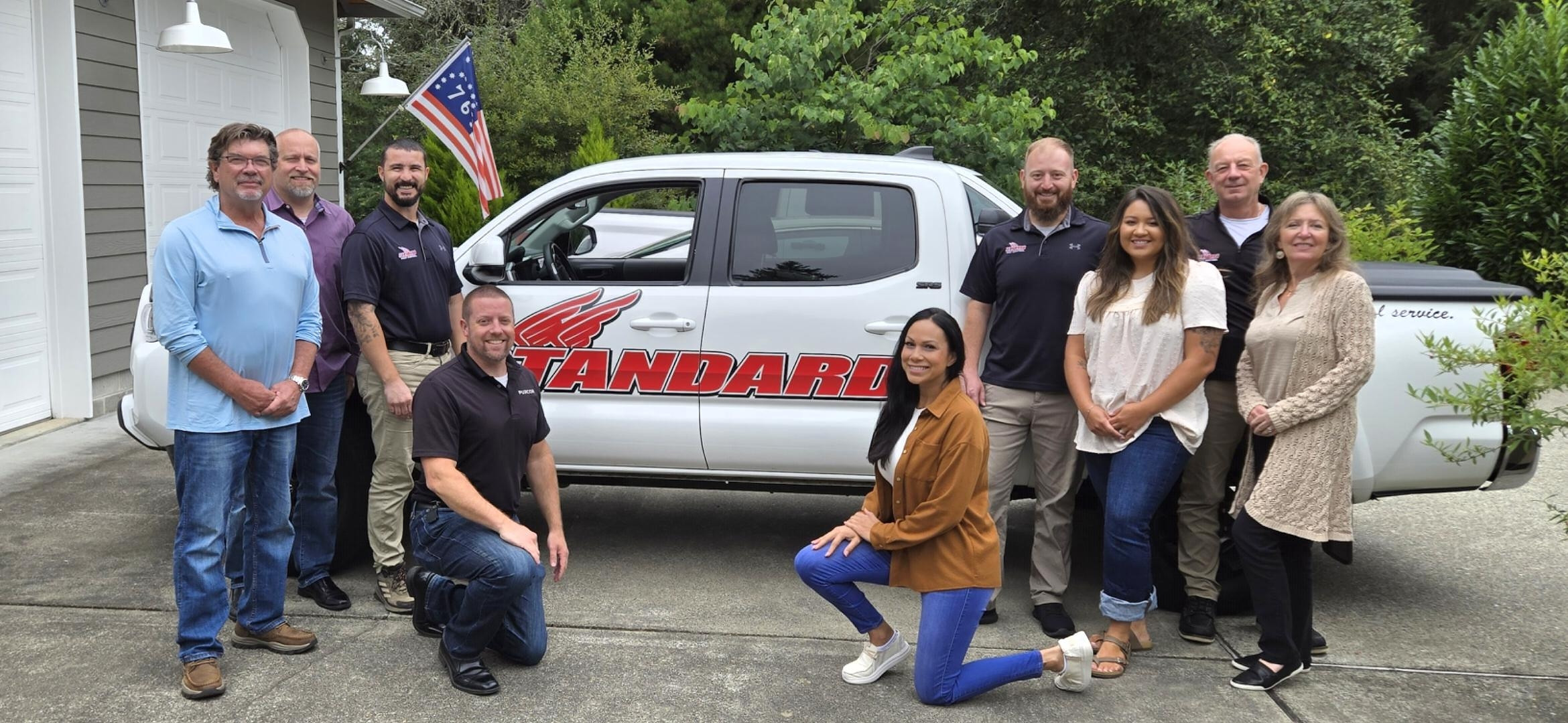 Ten adults pose for a group photo in front of a white pickup truck with “Standard” written on the side, representing Standard Pest Control. Some are standing while others kneel, with trees visible in the background, capturing a moment of camaraderie in Washington.