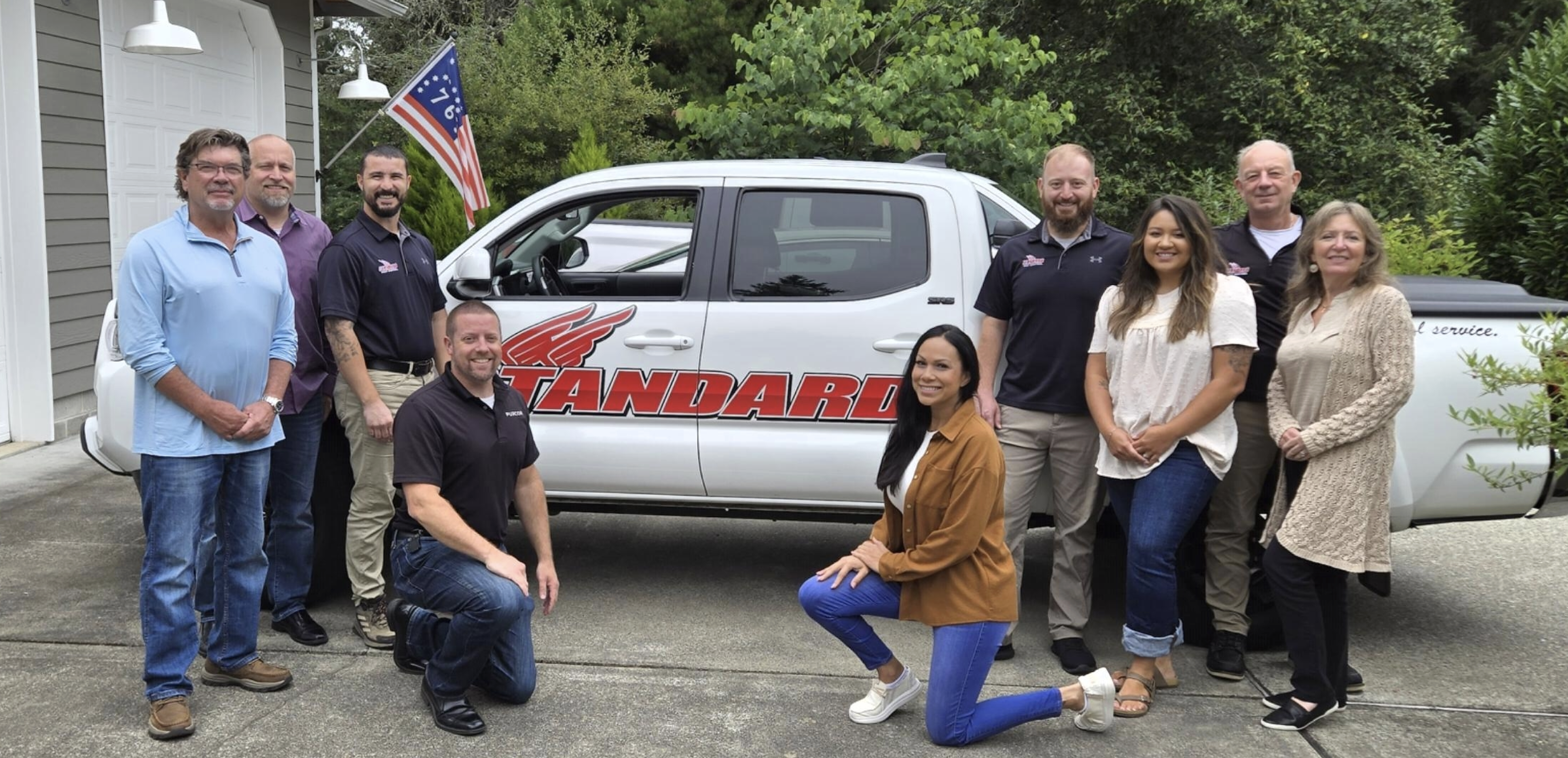 Eight people pose in front of a white pickup truck with the words “STANDARD PEST CONTROL” on the side. The group stands and kneels on a driveway next to trees and a house, with an American flag visible behind the truck, representing their commitment to quality pest control in Washington as part of the CERTUS family.